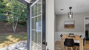 Un coin repas avec une table et des chaises en bois, un tableau accroché au mur et une vue sur l’extérieur grâce à une porte vitrée.