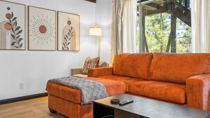 A living room with a sofa, ottoman, and a coffee table. There are three framed artworks on the wall, a lamp, and a window with curtains. - Luxury Studio Escape - Trails & Views (Sugarloaf)