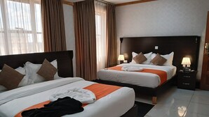 Habitación de hotel con dos camas, cada una con ropa blanca y almohadas marrones, cabecero negro, mesita de noche con lámpara y ventana con cortinas.