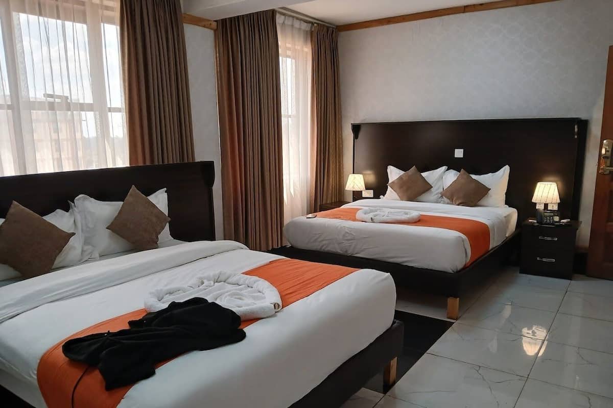 Camera d'albergo con due letti, biancheria da letto bianca e cuscini marroni, una testiera nera, un comodino con una lampada e una finestra con tende.