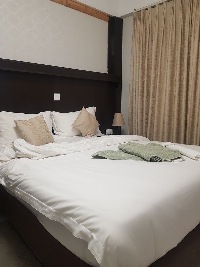 A neatly made bed with white linens and a green towel, a bedside lamp, and a window with beige curtains.