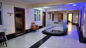 Un pasillo de hotel con piso de mármol, una zona de estar con una butaca naranja y una barandilla blanca alrededor de una columna central.