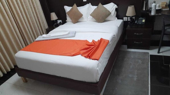 A hotel room with a bed, a nightstand, a lamp, a telephone, and a small table with a box and a bottle. - Grand Tenacity Hotel Kitale (Kitale)
