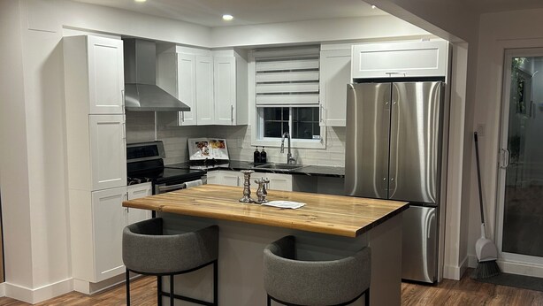 A modern kitchen with white cabinets, a stainless steel refrigerator, a wooden island with two bar stools, and a window with blinds. - Luxury sweet home (Longueuil)