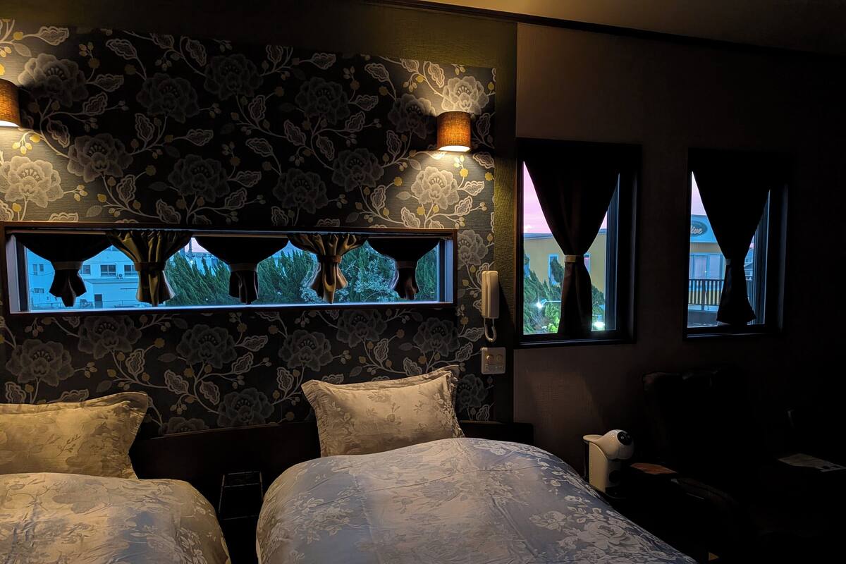 A hotel room with two beds, floral wallpaper, a window with curtains, and a wall-mounted phone.
