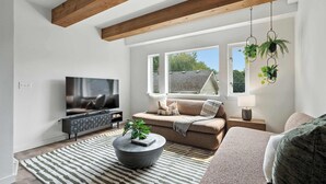 A modern living room with a large flat-screen TV, a sofa, a coffee table with a plant, and hanging plants. - The Adelynn 5BR - Sleek - Spacious - Walkable (Portland)