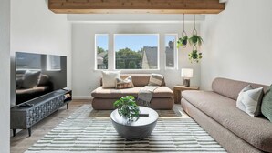 A modern living room with a sofa, TV, and coffee table, featuring a large window and hanging plants. - New Spanish-Style Townhome in Kerns | Walk to Bars & Cafes (Portland)