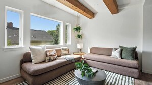 Un salón moderno con un sofá marrón, una mesa de centro redonda y una alfombra a rayas. Hay dos ventanas que ofrecen luz natural y vistas al exterior.