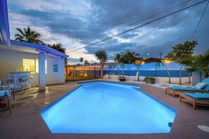 Una piscina con sillas de descanso, un edificio con puerta de vidrio y palmeras.