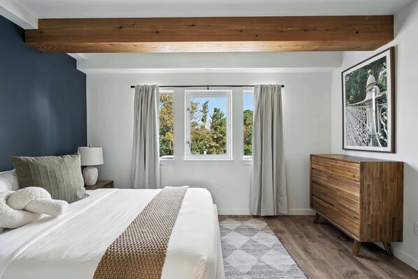 A bedroom with a large bed, a wooden dresser, a window with curtains, and a framed picture on the wall.