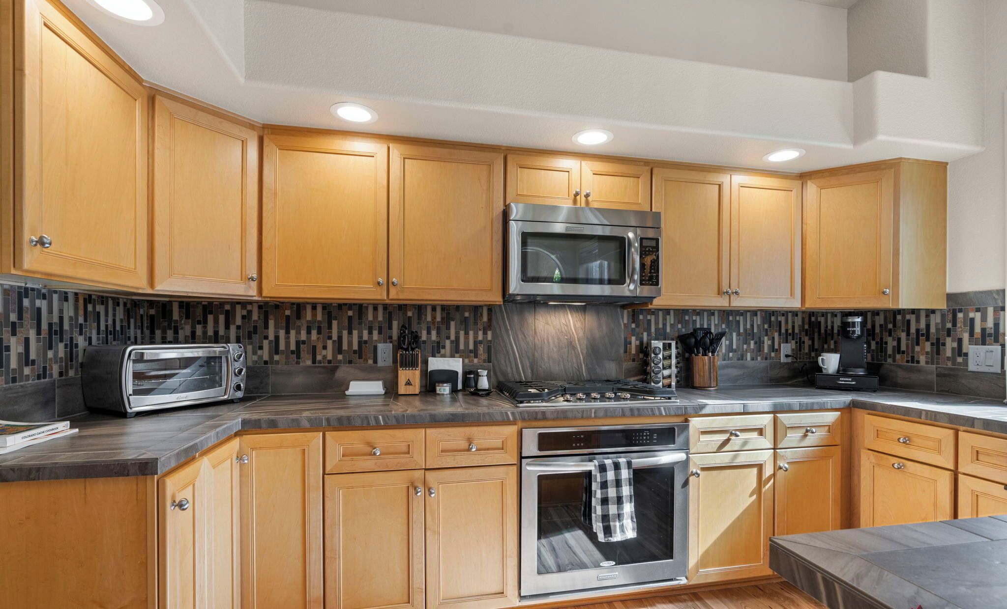 A modern kitchen with wooden cabinets, a stainless steel range, a microwave, a toaster, and a tiled backsplash.