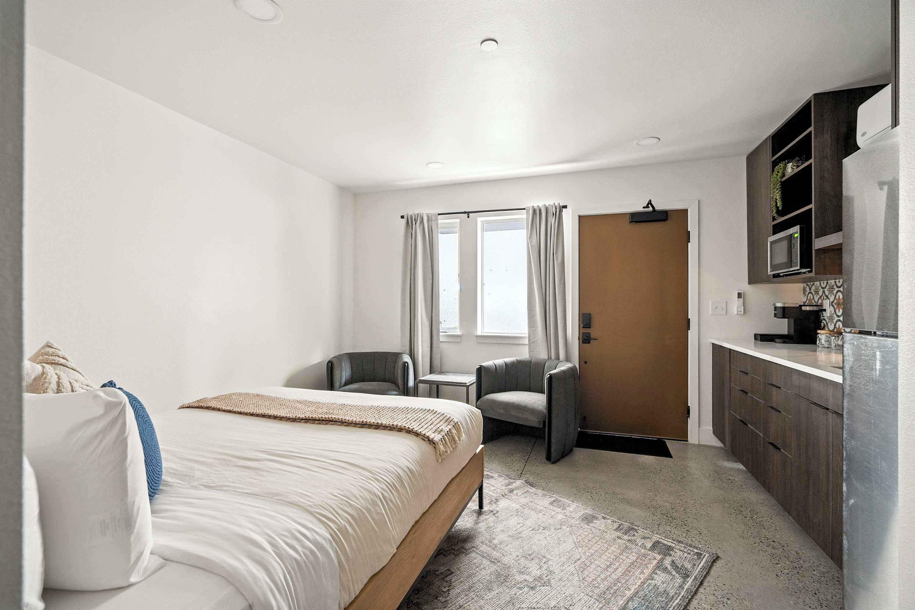 A hotel room with a bed, a desk, a chair, a kitchenette, and a door leading to another room.