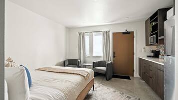 A hotel room with a bed, a desk, a chair, a kitchenette, and a door leading to another room.