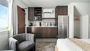 Une chambre d’hôtel compacte comprenant un coin cuisine, un fauteuil gris, un lit avec une couverture à motifs et une vue sur l’extérieur.