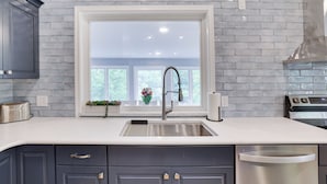 A kitchen with a white countertop, dark cabinets, a stainless steel sink, and a window with a view of greenery outside.