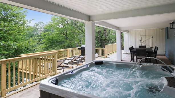 A hot tub on a covered deck with a wooden railing, overlooking a body of water and surrounded by greenery.