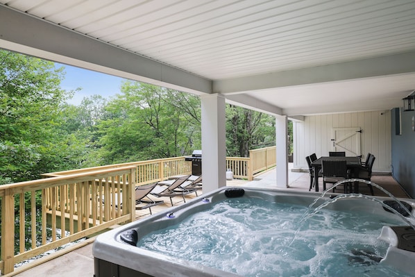 Un jacuzzi en un deck techado con barandilla de madera, con vista a un cuerpo de agua rodeado de vegetación.