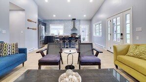 A modern living room with a blue sofa, yellow sofa, and a coffee table with decorative items. There are two armchairs and a dining table in the center. The kitchen is visible in the background with a large island and bar stools.