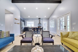 Una sala de estar moderna con un sofá azul, un sofá amarillo y una mesa de centro con objetos decorativos. Hay dos sillones y una mesa de comedor en el centro. Se ve la cocina al fondo, con una isla grande y taburetes de bar.