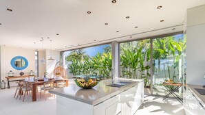 A modern kitchen with a central island, large windows, and a dining area with a round mirror and hanging light fixtures.