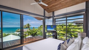 A bedroom with a large bed, a TV on a stand, and a view of the ocean and palm trees.