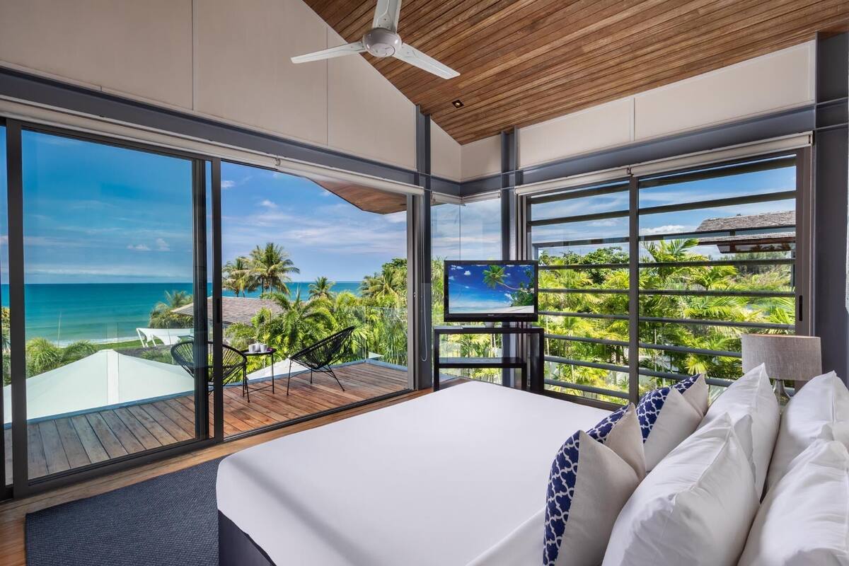 A bedroom with a large bed, a TV on a stand, and a view of the ocean and palm trees.