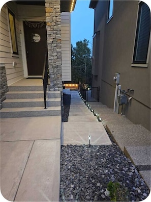 A residential property with a stone pillar, a front door with a sign, a concrete pathway, and a small garden area with rocks and a plant.