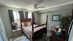 Une chambre à coucher avec un lit à baldaquin, un ventilateur de plafond, une fenêtre avec des rideaux, une table de chevet avec une lampe, une chaise, une commode et une plante.