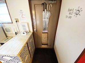 A narrow hallway with a wooden counter on the left, a door with a coat rack and umbrellas, and a wall with a 'Have a nice day' sign. - Nostalgic Japan! Direct to Shinjuku & Shibuya on JR Station. (Kita City, Ukima)