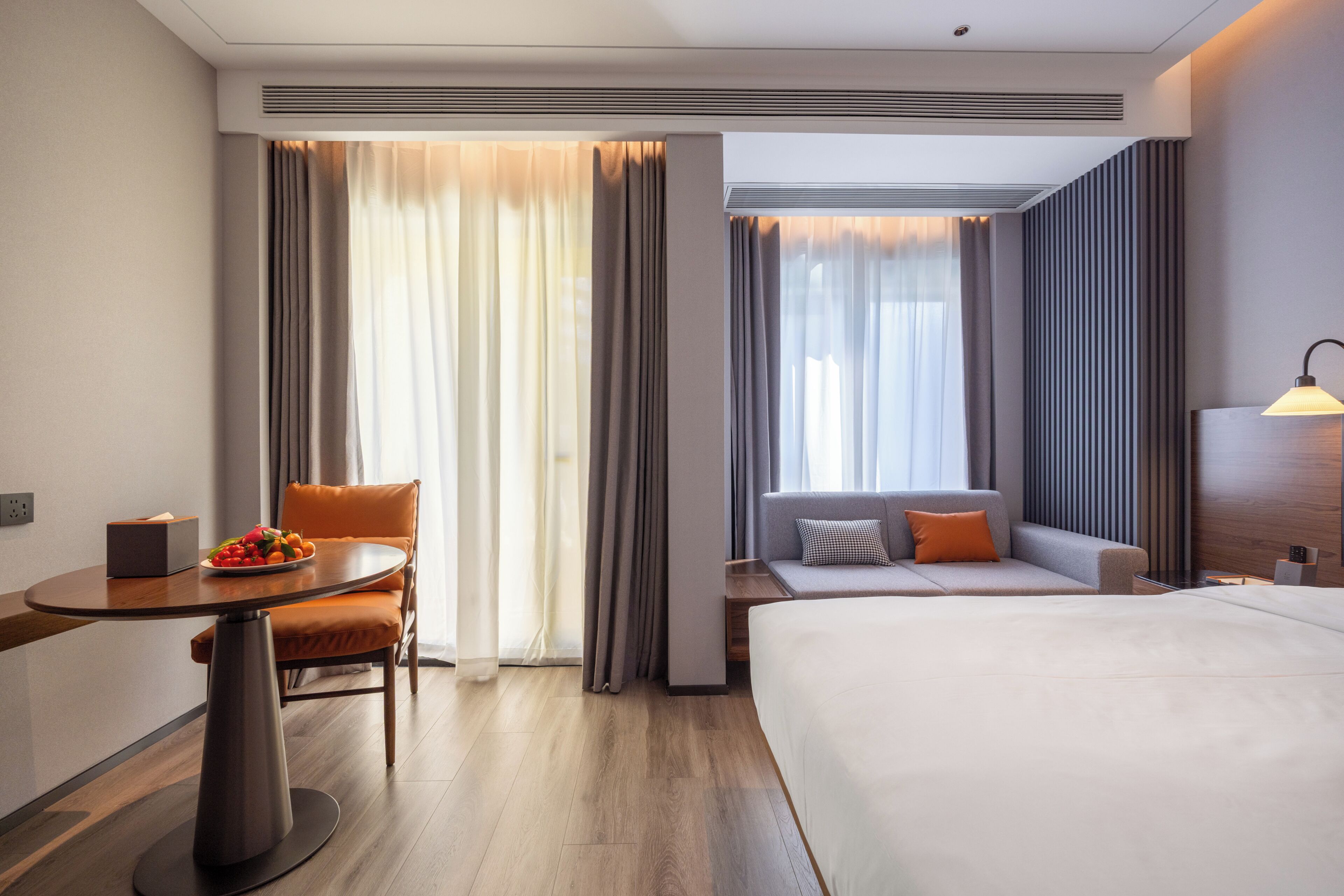 A hotel room with a bed, a sofa, a table with a fruit bowl, and a desk with a lamp.