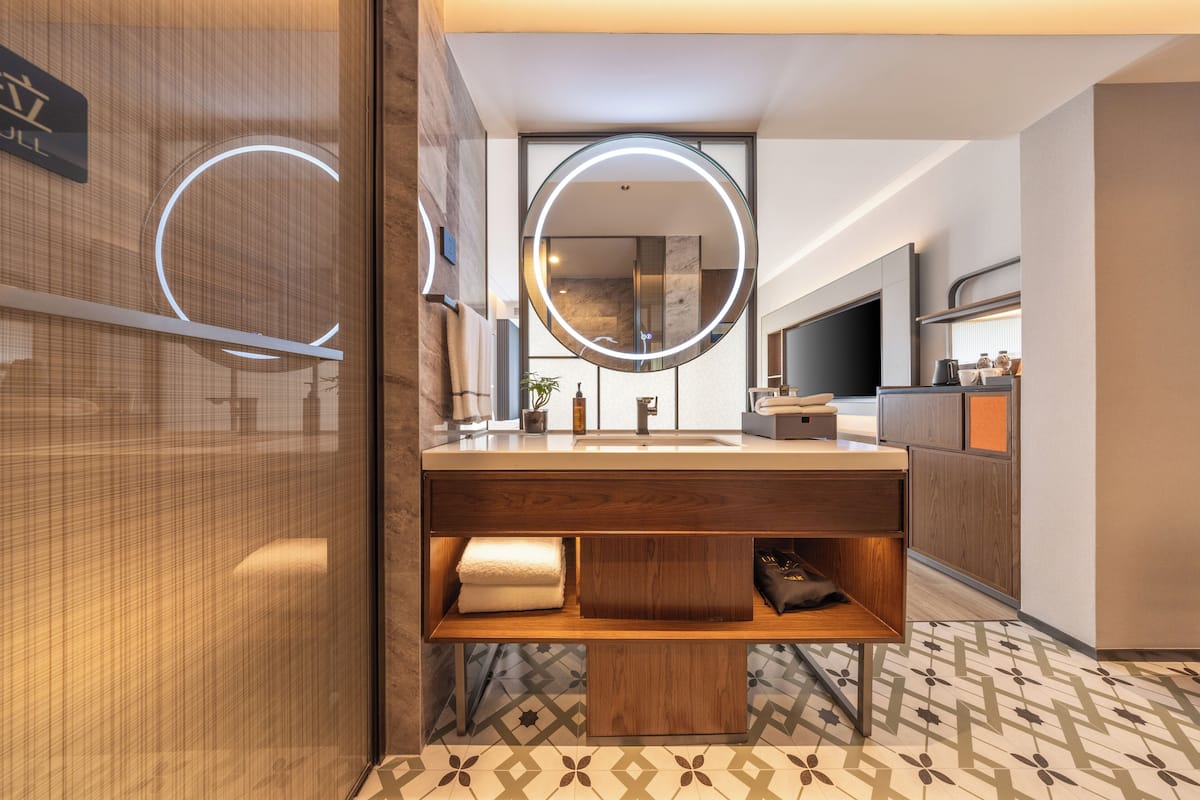 Une salle de bain moderne avec un meuble-lavabo en bois, un grand miroir avec un éclairage circulaire et un sol carrelé.