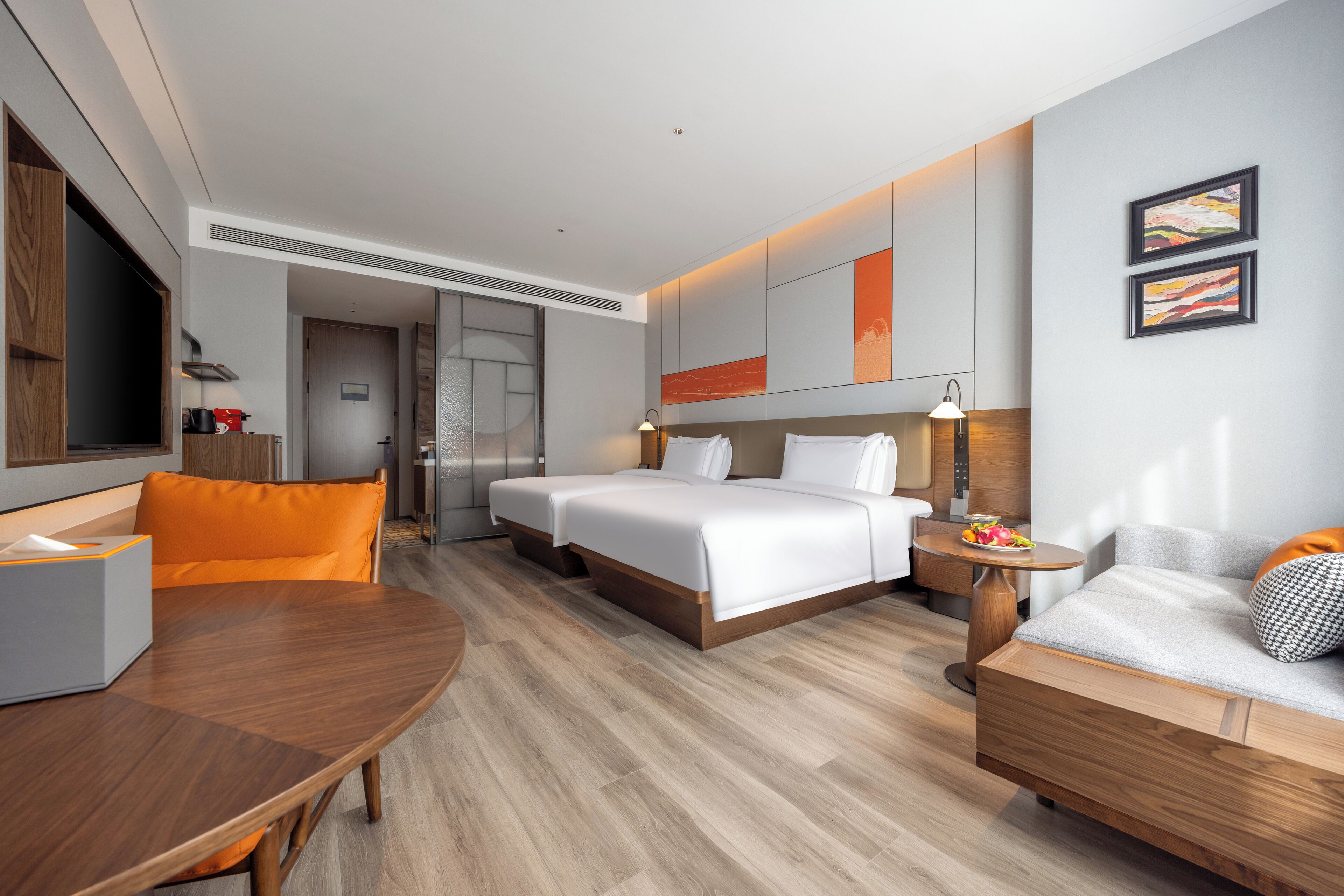 Ein modernes Hotelzimmer mit Holzboden, einem Bett mit weißen Leinen, einem Sofa mit orangefarbenen Kissen, einem kleinen Tisch mit Obstschale und einem Schreibtisch mit Lampe.