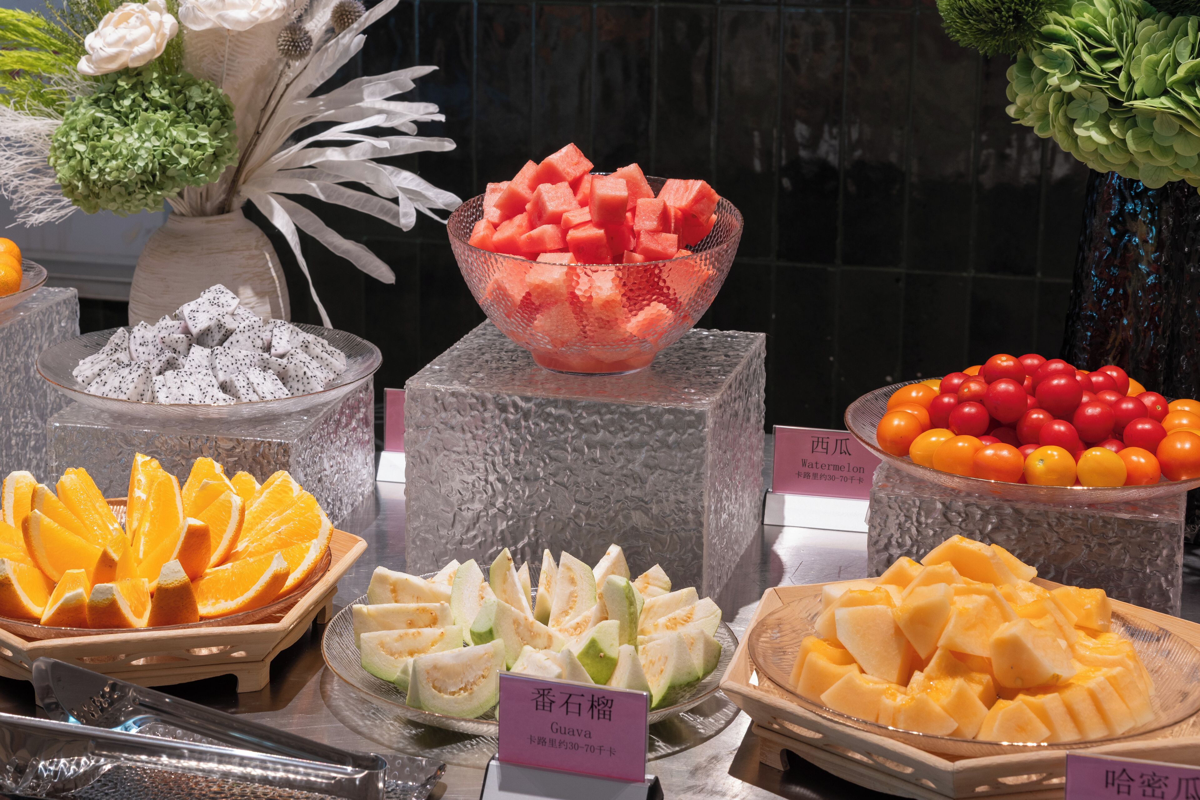 A fruit display with various types of fruits in bowls and plates, including watermelon, oranges, and tomatoes.