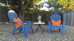 Due poltrone Adirondack blu con cuscini arancioni, un tavolino rotondo con un vaso bianco e un albero sullo sfondo.