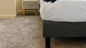 Un lit bien fait, avec des oreillers et une couverture, une table de chevet avec une lampe et une petite plante, et une fenêtre avec des stores.