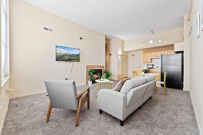 Uma sala de estar moderna com lareira, sofá, mesa de jantar e área de cozinha com armários de madeira e geladeira preta.