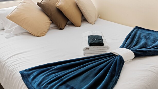 Una cama bien hecha con una manta azul, varios almohadones y una toalla doblada con una etiqueta.