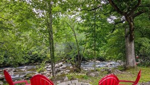 Un campamento con dos sillas rojas y un brasero rodeado de un bosque y un arroyo.
