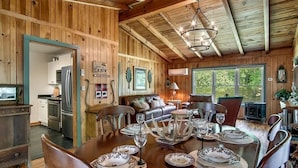 Interior de una cabaña de madera con una mesa de comedor preparada para cuatro personas, una cocina equipada con electrodomésticos modernos y una zona de estar acogedora con chimenea.