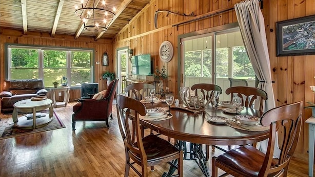 Interior de una cabaña de madera con una mesa de comedor para seis personas, rodeada de sillas de madera. Hay un sofá y un televisor en la sala.
