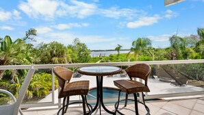 Un balcon agrémenté de fauteuils en osier et d’une table en verre, offrant une vue sur une piscine et une végétation luxuriante.