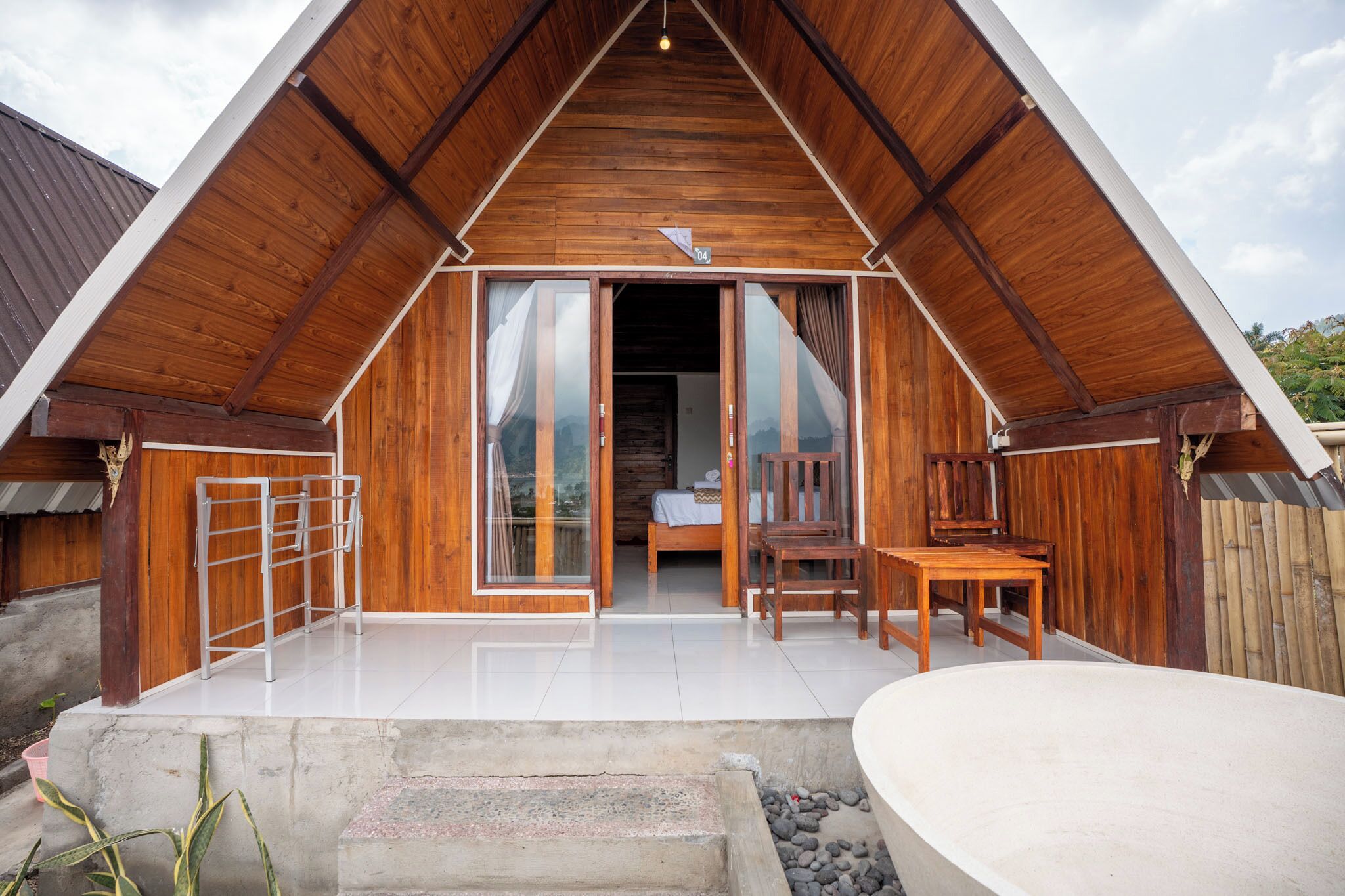 Una cabaña de madera con techo inclinado, puerta de vidrio, un pequeño patio con mesa y sillas, y una plataforma de concreto con una bañera blanca.