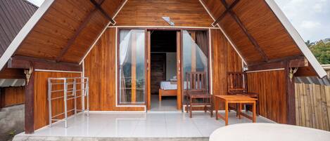 Una cabaña de madera con techo inclinado, puerta de vidrio, un pequeño patio con mesa y sillas, y una plataforma de concreto con una bañera blanca.