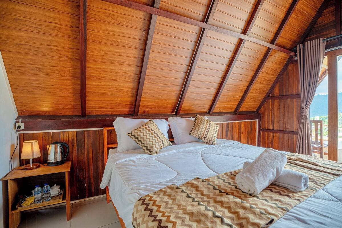 A wooden ceiling, a bed with pillows, a nightstand with a lamp, and a view of the outdoors.