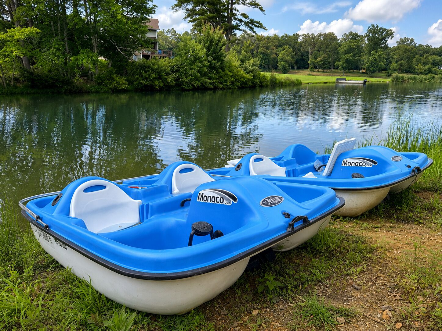 Two blue and white Monaco brand inflatable boats parked by a calm lake with lush greenery and a bench in the background.