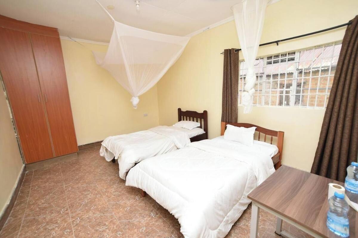A room with two beds, a wooden wardrobe, a table with water bottles, and a window with curtains.