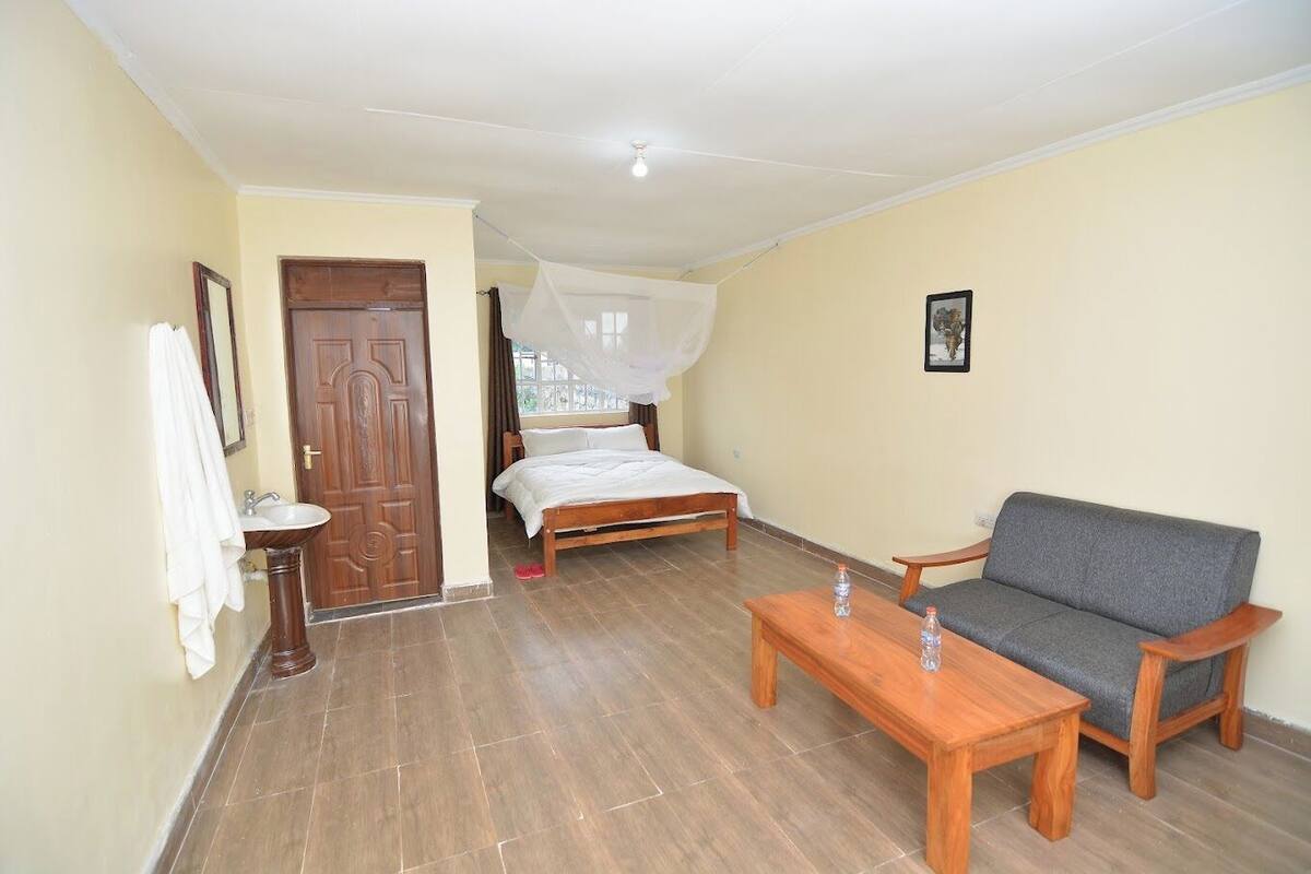 A room with a bed, a sofa, a wooden table, and a bathroom with a sink and a door.