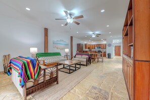 A spacious living area with a sofa, chairs, a coffee table, and a dining table. There is a kitchen in the background with wooden cabinets and a microwave.