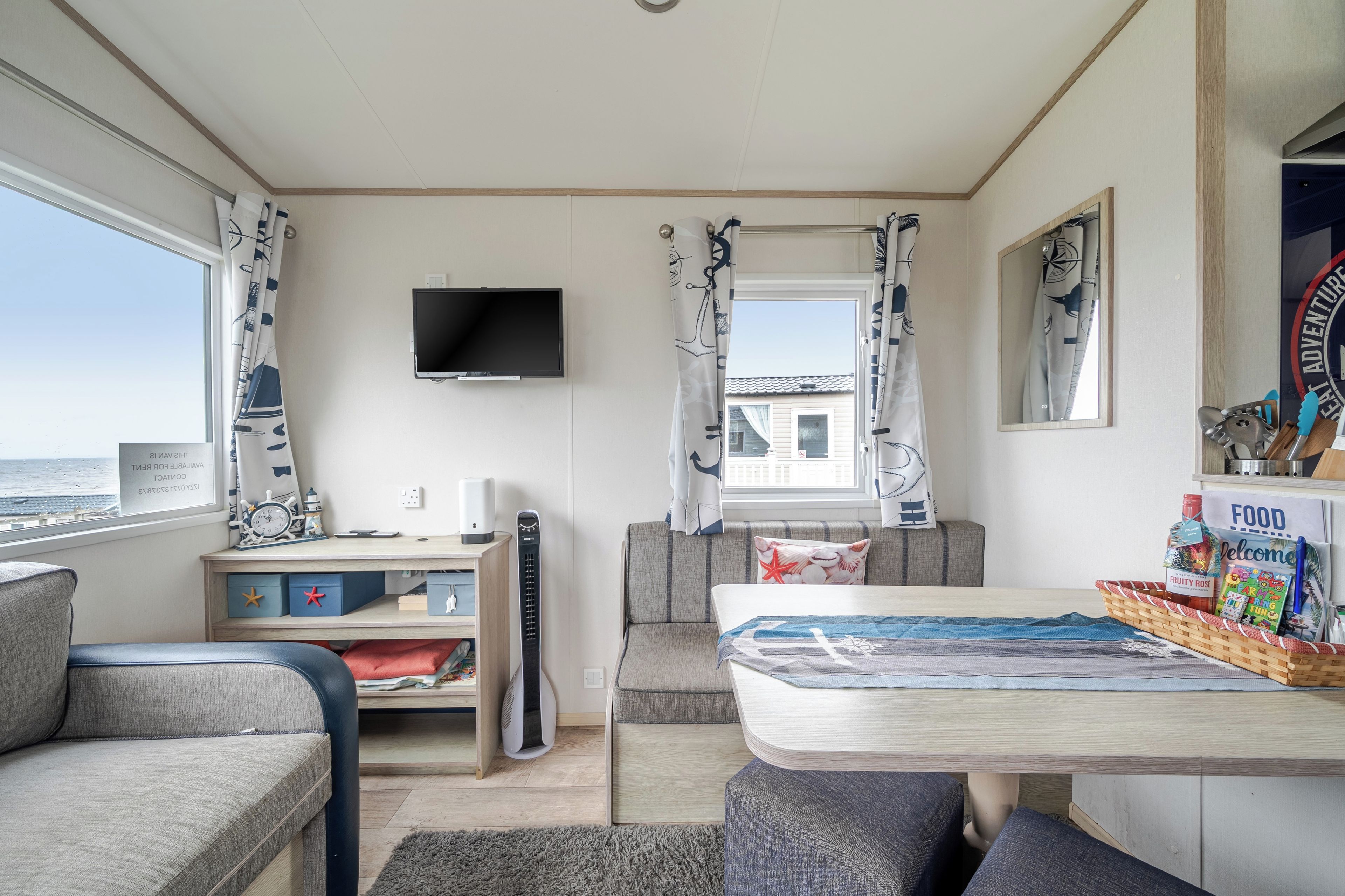 Een kamer met een televisie, een tafel met een bord waarop 'FOOD' staat, een raam met gordijnen en uitzicht op zee.
