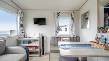 Een kamer met een televisie, een tafel met een bord waarop 'FOOD' staat, een raam met gordijnen en uitzicht op zee.
